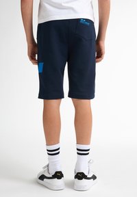 Námořnické šortky vyrobené z bavlněné směsi, s modrou nášivkou na levé noze a značkovou visačkou na zadní straně. Spárovány s bílými proužkovanými ponožkami a teniskami.