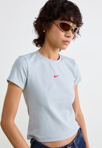 Kvinde med kort krøllet hår iført olivengrønne katteøje-solbriller, lyseblå Nike t-shirt og mørkeblå jeans foran en ensfarvet hvid baggrund.