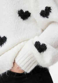 Miękka, biała dzianinowa sweter z czarnymi wzorami serc noszony przez osobę z rękami schowanymi w kieszeni czarnych spodni.