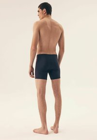 Granatowe kąpielówki o dopasowanym kroju, z elastycznym pasem i detalami stębnowania z boku. Model stoi na boso i odwraca się nieco bokiem.