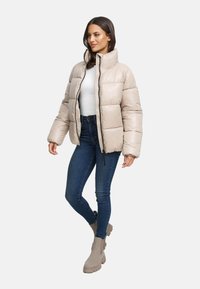 Giacca puffer beige con colletto alto, design trapuntato, chiusura con zip, abbinata a jeans scuri e stivaletti beige. Texture liscia.