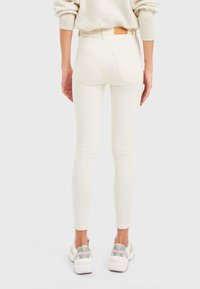 Jeans blancos ajustados con cintura alta, dos bolsillos traseros y detalles de costura mínimos, combinados con zapatillas grises y verdes.
