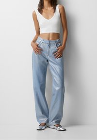 Jeans de mezclilla anchos de un azul claro, con cintura alta y hardware mínimo, combinados con un top ajustado blanco corto y flats plateados.