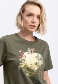 T-shirt in cotone verde oliva con scollatura rotonda, caratterizzata da una stampa grafica dettagliata di una scena bizzarra con vari elementi giocosi.