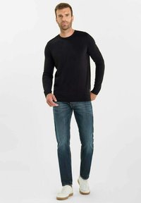 Uomo che indossa una maglietta nera a maniche lunghe, jeans blu e sneakers bianche, in piedi contro uno sfondo bianco.