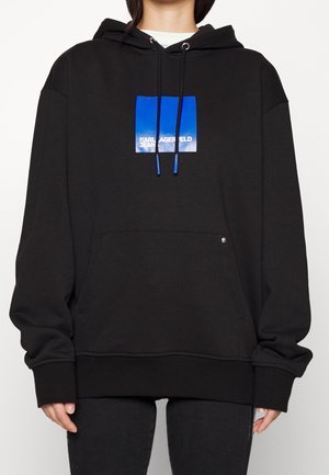 Osoba nosząca czarną, oversize’ową bluzę z kapturem z przednią kieszenią oraz niebieskim kwadratowym logo z białym napisem „KARL LAGERFELD JEANS” na piersi.