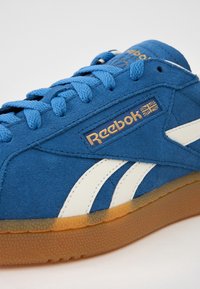 Blå mockasinsneaker med vita sidostrippar, strukturerad ovandel, vadderad plös, gulddetalj på logotypen och en genomskinlig sula i gummimaterial.