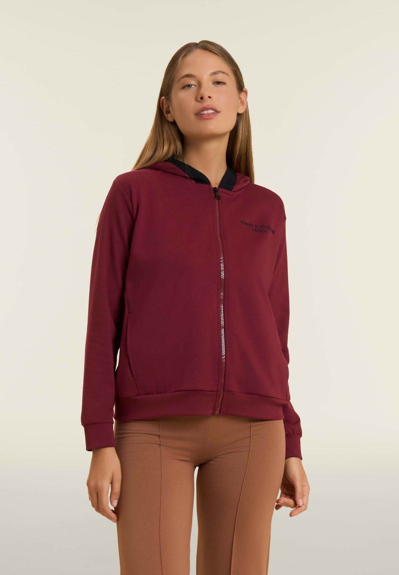 Sudadera con cremallera color burdeos y capucha forrada en negro, con bolsillos delanteros y un logo discreto en el lado izquierdo del pecho, además de puños acanalados.