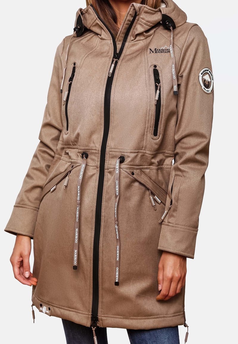 Femme portant un parka beige à capuche avec fermeture éclair à l’avant, poches zippées poitrine et taille, logo sur la poitrine et écusson sur le bras, et cordons de serrage ajustables à la taille.