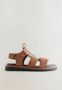 Sandal de ante marrón con tres tiras anchas, soporte de talón trasero y suela de goma negra con un acento marrón en el borde.