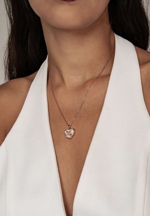 Lessing Herzanhänger aus roségold mit filigranen Blumenmotiven an einer zarten Kette, getragen zu einem tief ausgeschnittenen weißen Kleidungsstück.