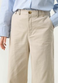 Beige cotton trousers with a straight-leg cut, featuring a button closure, side pockets, and soft texture. Blue-striped collared shirt worn.