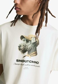 T-shirt crema con una grafica di un cane con cappello e sciarpa. Il testo recita "SNOUTCHIC" sopra e "Look deciso con un lato selvaggio" sotto.