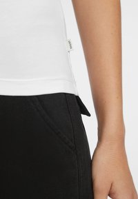 Weißes Baumwoll-Top mit einem Seitennähten-Logosticker, kombiniert mit schwarzen Jeans mit einer dezenten Tasche und sichtbaren Nähdetails.