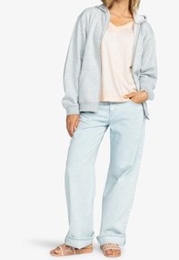 Personne portant un sweat à capuche zippé gris clair, un t-shirt rose pâle, un jean bleu clair retroussé et des sandales beige à enfiler avec des lettres.