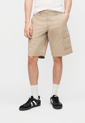 Mann, der beige Cargoshorts, weiße Socken und schwarze Turnschuhe mit weißen Streifen trägt, vor einem schlichten weißen Hintergrund steht.