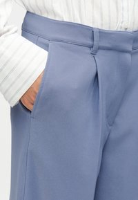 Gaiši zilas bikses ar gludu faktūru, priekšpusē ar krokām, sānu kabatām un piegrieztu vidukļa jostu. Redzamas svītrainas kreklu aproces.