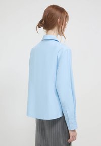 Camicia azzurra chiaro in cotone; presenta un colletto, maniche lunghe e una vestibilità rilassata. Leggero lucido e texture morbida.
