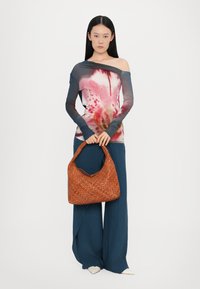 Langärmliges, schulterfreies Oberteil mit floralem Design in verschiedenen Rosatönen und Blau; kombiniert mit weiten, dunkelblauen Hosen und einer geflochtenen braunen Handtasche.