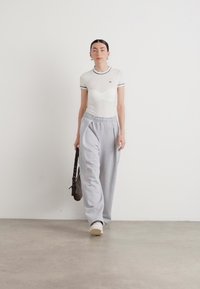 Weißes, tailliertes T-Shirt mit schwarzem Rand, kombiniert mit grauen Jogginghosen. Das Modell trägt eine schwarze Tasche und weiße Sneakers.