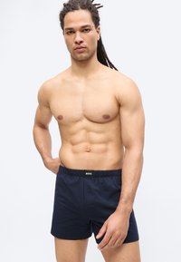 Homme musclé avec des dreadlocks portant un boxer BOSS bleu foncé, debout devant un fond clair uni.