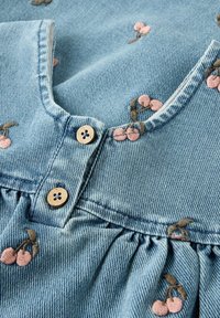 Vaaleansinistä farkkukangasta, jossa on vaaleanpunaisia kirjailtuja kirsikkakuvioita, poimutettuja yksityiskohtia ja kaksi pientä puista nappia V-pääntiellä.