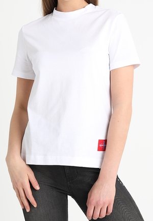 Kvinde iført plain hvid kortærmet T-shirt med rød Calvin Klein logo-patch nær kanten, kombineret med sorte jeans.