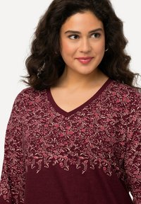 Burgunderfarbenes Longsleeve-Shirt mit V-Ausschnitt. Ausgestattet mit einem floralen Muster in Pink und Beige, weichem Material und einem lässigen Schnitt.
