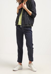 Black zip-up bomber jacket, light green top, navy tapered pants, and white sneakers. Smooth textures and functional zippers on the jacket.