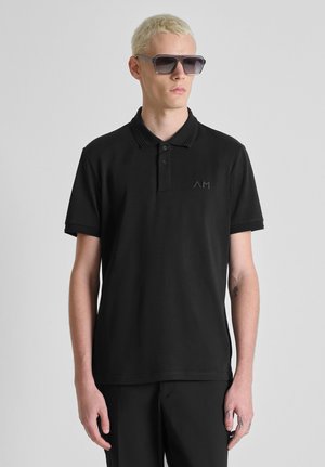 Hombre rubio que lleva gafas de sol oscuras, una camiseta tipo polo de manga corta negra con un sutil logo "AM" y pantalones negros sobre un fondo gris.
