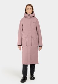 Lungo impermeabile rosa con cappuccio, due tasche frontali e chiusura a zip. Indossato con pantaloni neri e stivali neri massicci.