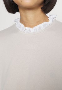 Helles beiges Oberteil aus strukturiertem Stoff mit einem weißen Rüschensaum-Kragen. Der Ausschnitt ist rund und glatt.