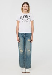 T-shirt bianca a girocollo con grafica nera "NEW YORK", abbinata a jeans larghi strappati di un azzurro chiaro e sneakers bianche. Stile semplice e casual.