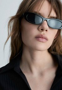 Ochelari de soare negri, de formă ovală, cu ramă groasă, textură netedă și lentile reflective, asortați cu o cămașă neagră cu dungi fine.