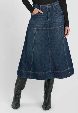 Femme portant une jupe midi en jean bleu foncé en ligne A avec des poches, associée à des bottes noires montantes et un haut à manches longues noir.