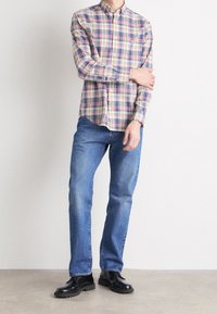 Man staat, droeg een veelkleurig geruit overhemd met lange mouwen, blauwe spijkerbroek en zwarte gepolijste schoenen, tegen een witte achtergrond.