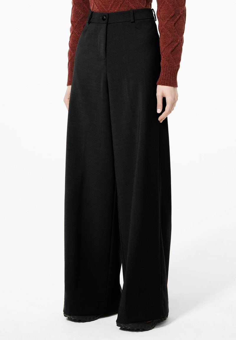 Vrouw met hoge taille, wijde zwarte broek en een roestkleurige, lange mouwen, gestructureerde trui, staand tegen een witte achtergrond.