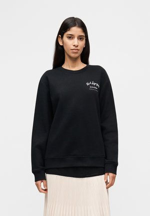 Frau mit langen dunklen Haaren, die ein schwarzes Sweatshirt mit rundem Ausschnitt und weißem Schriftzug trägt, dazu einen hellen plissierten Rock. Sie steht vor einem weißen Hintergrund.