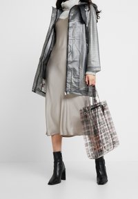 Transparentní pláštěnka se vzorem v šedé a černé barvě, vrstvená přes béžové šaty. Celý outfit doplňuje průhledná kostkovaná taška a černé kotníkové boty.