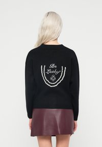 Melna džemperis ar brīvu piegriezumu, kas rotāts ar baltām pērlītēm, veidojot dekoratīvu rakstu un tekstu, apvienots ar bordo ādas svārkiem.