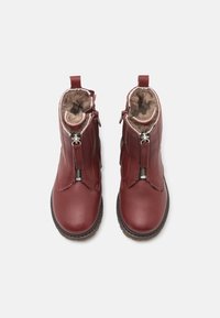 Botas de tobillo de cuero rojo con cremalleras, forro de pelo sintético, diseño curvado y suelas de goma marrón. Presentan costuras y lengüetas para ponerlas y quitarlas fácilmente.
