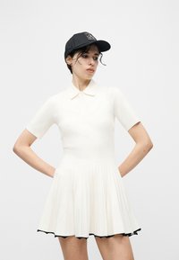 Γυναίκα που φοράει λευκό πλισέ φόρεμα με κοντά μανίκια και γιακά, και μαύρο καπέλο μπάσκετ, ποζάρει με τα χέρια στη μέση μπροστά από απλό φόντο.