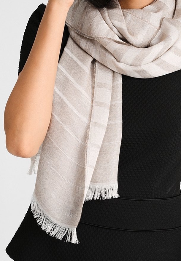 Femme portant une écharpe rayée beige clair avec des franges sur un haut noir texturé, tenant l'écharpe près de son visage.