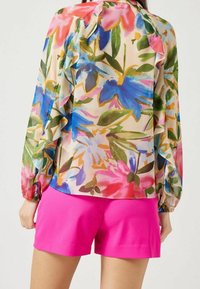 Blusa floreale con maniche lunghe a sbuffo in tonalità di blu, rosa e verde; dettagli arricciati lungo la schiena. Abbinata a pantaloni corti rosa.