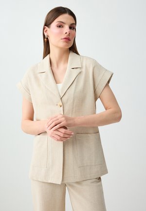 Femme debout devant un fond uni, portant un blazer beige à manches courtes et un pantalon assorti, les mains jointes à la taille, regardant devant elle.