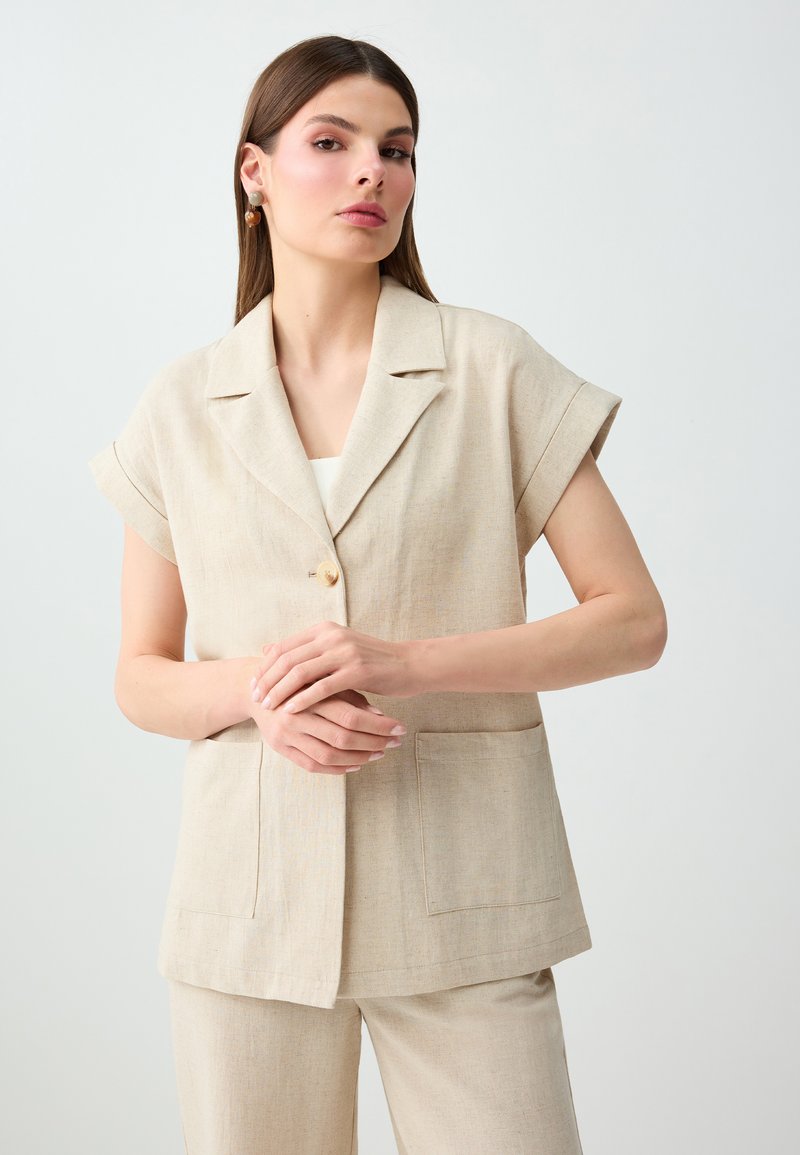 Femme debout devant un fond uni, portant un blazer beige à manches courtes et un pantalon assorti, les mains jointes à la taille, regardant devant elle.