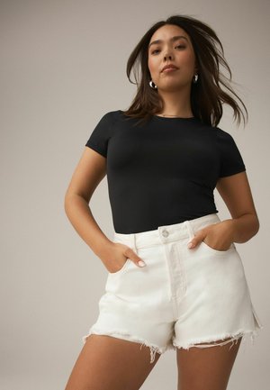 Tânără cu păr lung poartă un tricou negru mulat și pantaloni scurți albi din denim, cu talie înaltă și tivuri ciufulite, cu mâinile în buzunare.