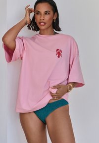 Lyserød oversized t-shirt med en rød hummerekspedition på brystet. Model iført teal teksturerede bikini-bund, stående foran en hvid væg.
