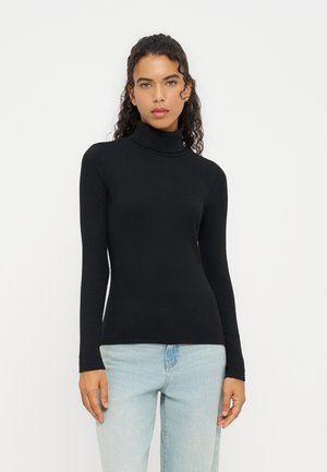Femme aux cheveux bouclés portant un col roulé noir moulant et un jean bleu clair, debout devant un fond blanc uni.