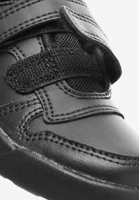 Chaussure en cuir noir avec une surface texturée, dotée d'une grande sangle en Velcro noire et de coutures contrastées. Bout arrondi et semelle rembourrée.